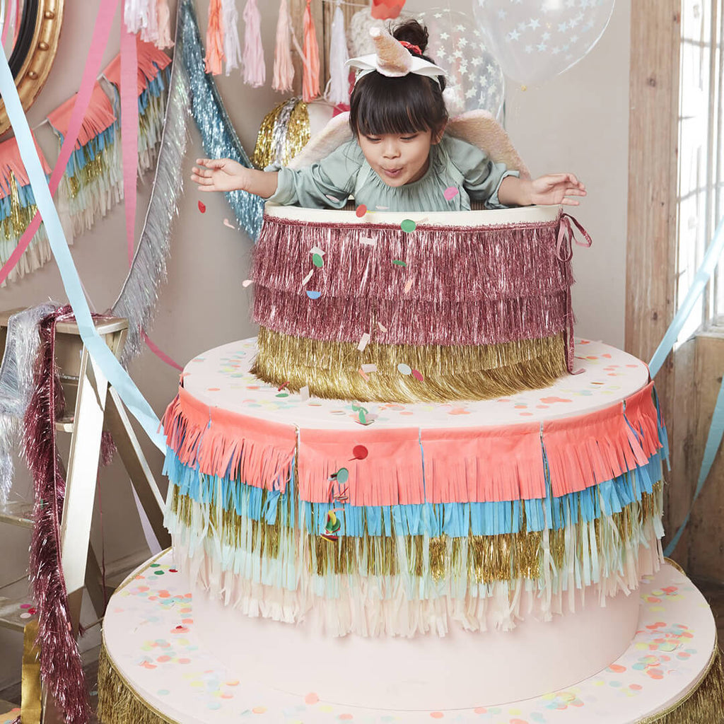 meri-meri-party-pink-gold-tinsel-fringe-garland-banner-styled-with-child-coming-out-of-giant-birthday-cake-prop
